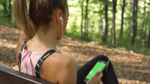 Девочка отдыхает на деревянной скамейке в лесу после долгого бега и слушает музыку в наушниках — стоковое видео