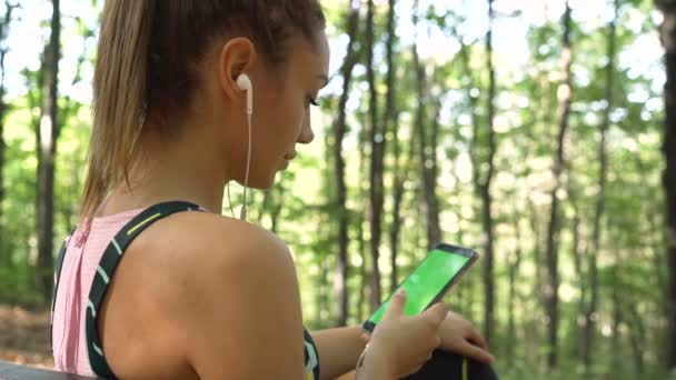 Красива дівчина в спортивному одязі сидить на лавці в лісі, розслабляє і слухає музику з телефону — стокове відео
