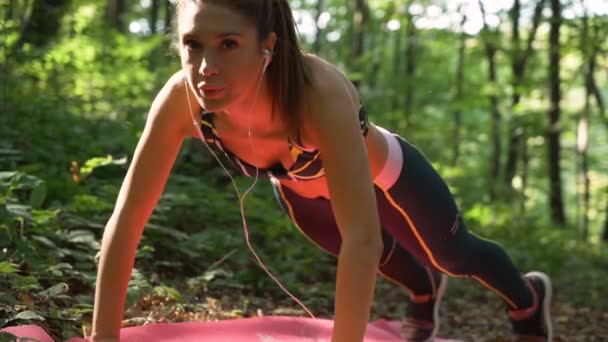 Vista cercana de la chica en forma con auriculares haciendo flexiones en la esterilla de yoga en el parque — Vídeo de stock