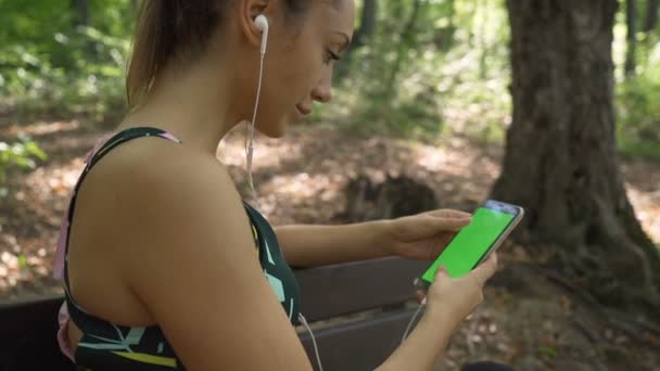 Продумана активна дівчина сидить на лавці в парку, слухаючи музику зі смартфона — стокове відео