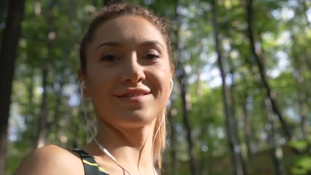 Nízký úhel pohledu brunetky dívka s culíkem, poslech hudby a úsměv na kameru s rozmazaným pozadím — Stock video