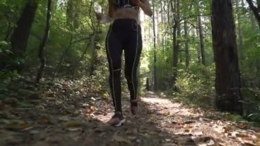 Spor kıyafetli bir bayan koşucu ve spor ayakkabılarıyla ormanda koşuyor, elinde su şişesi var.