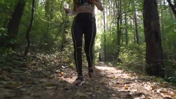 Corredor femenino en traje deportivo y zapatillas deportivas corriendo por un sendero forestal, llevando botella de agua — Vídeos de Stock