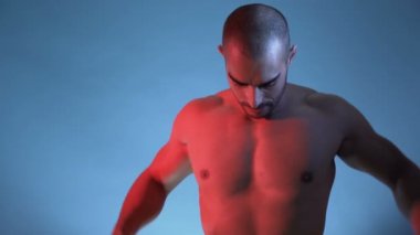 Ton halinde ve kaslı kaslı adam mavi bir arka planın önünde duruyor..