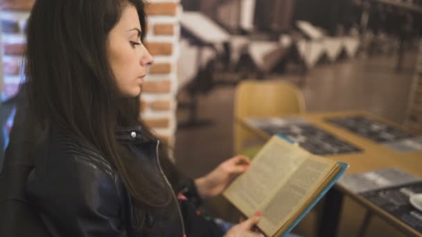 Молодая женщина с каштановыми волосами и кожаной курткой читает книгу с голубыми обложками в книжном кафе или кафе — стоковое видео