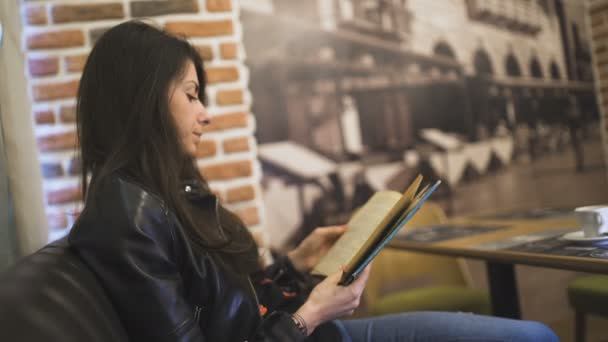 Mujer joven concentrada en jeans y chaqueta de cuero negro libro de lectura en la cafetería — Vídeos de Stock