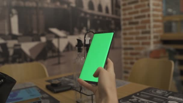 Oigenkännlig kvinna som använder smartphone med grön skärm sitter på ett café och leker med håret — Stockvideo