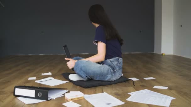 Schöne junge Frau mit Brille sitzt auf Holzboden, arbeitet am Laptop, schreibt etwas in ihr Notizbuch, Home Office. — Stockvideo