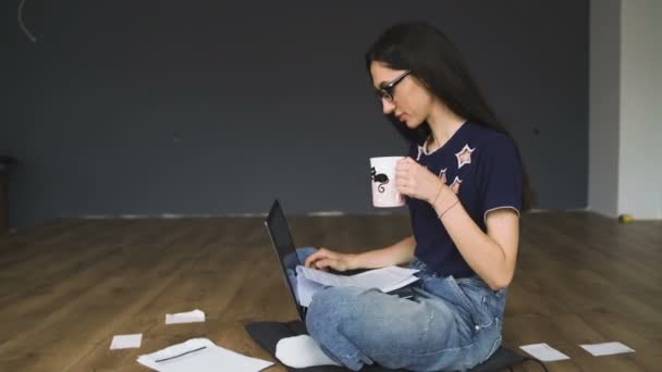 Mujer con el pelo largo y negro y gafas tomando un sorbo de café mientras trabaja desde casa, sentada en el suelo de madera con papeles a su alrededor — Vídeos de Stock