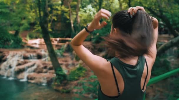 Mujer en forma arreglando su cabello mientras disfruta de un día soleado en el bosque, viendo las cascadas del bosque — Vídeo de stock