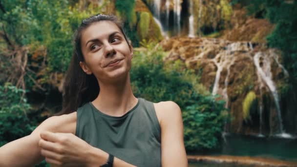 Позитивна усміхнена молода жінка бере селфі перед великим красивим лісовим водоспадом, що бризкає вниз у ставку — стокове відео