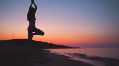 Genç fit kadın silueti gün batımında deniz kumsalında yoga yapıyor.