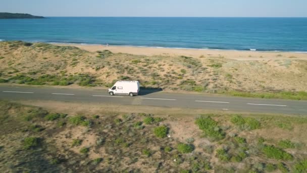 Дрон йде за білим фургоном або мінібусом вздовж прибережної дороги біля піщаного пляжу. — стокове відео