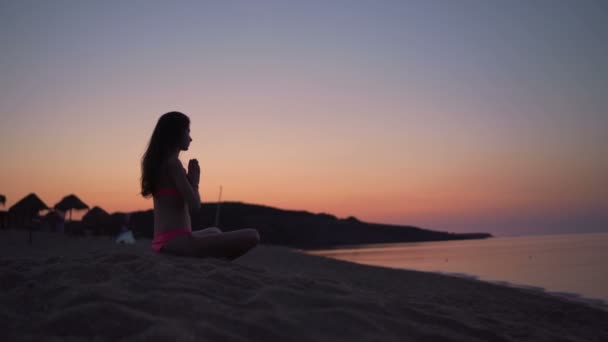 Профіль жінки в бікіні Молитва, що сидить на піщаному пляжі під час заходу сонця — стокове відео