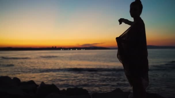 Silueta de mujer cubierta de bufanda apuntando al horizonte del mar con dramático cielo al atardecer sobre el mar — Vídeos de Stock