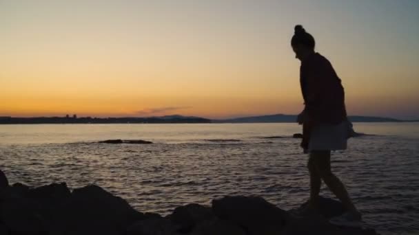 Young woman walking on rocky beach against sunset sky and reflecting sea surface — Stock Video