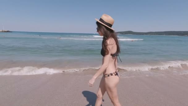Güneş şapkalı ve bikinili atletik bir kadın kumsalda çıplak ayakla yürüyor ve denize giriyor. — Stok video