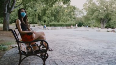 Yüz maskeli yalnız bir kadın birini bekliyor, saatini kontrol ediyor. Parktaki bankta yalnız bir kadın oturuyor.