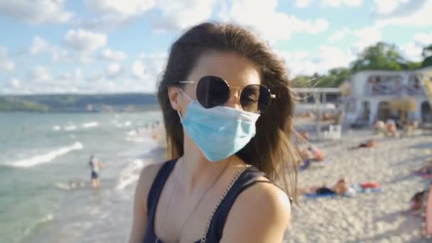 Mujer morena con gafas de sol y máscara médica protectora mirando el mar en la playa — Vídeo de stock