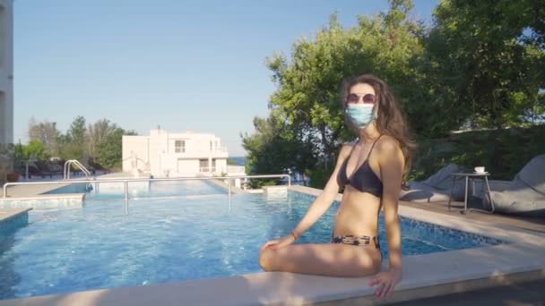 Mujer en bikini negro con mascarilla quirúrgica azul posando en la piscina en un caluroso día de verano — Vídeo de stock
