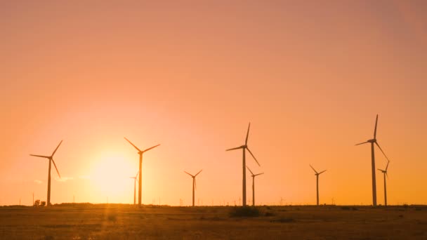 Draaiende windturbines in het veld tegen de grote zon bij zonsondergang met warme oranje lucht. Eco-energieconcept — Stockvideo