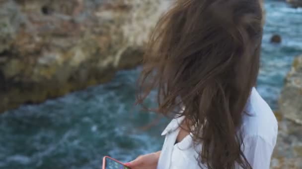 Hermosa joven con el pelo soplado por el viento, mirando el mar sentado en el borde del acantilado — Vídeo de stock