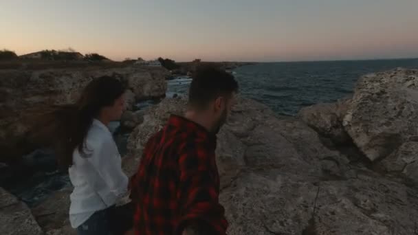 Jovem casal caminhando na costa rochosa atingindo a borda com o horizonte do mar calmo ao pôr do sol em frente — Vídeo de Stock