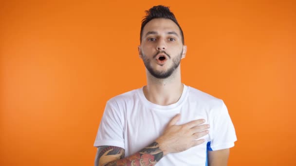 Joven fanático del fútbol cantando himno con la mano derecha en su corazón. Aislado sobre fondo naranja. Concepto patriotismo — Vídeos de Stock