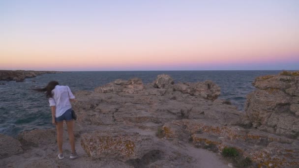Красива молода жінка з волоссям, підірваним вітром, дивиться на захід сонця з краю каменю — стокове відео
