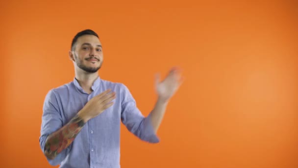 Fiatal mosolygós férfi táncol, és bemutatja oldalra a kezét, elszigetelt narancs háttér — Stock videók