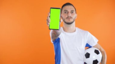 Futbol fanatiği beyaz mavi gömlekli ve toplu yeşil ekran akıllı telefon arkaplanı gösteriyor. 