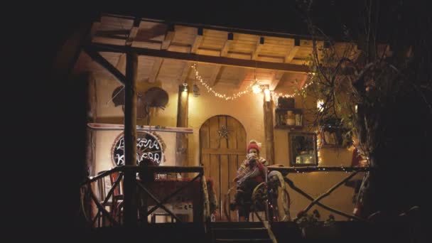 Wide view of pregnant woman drinking tea and swings in a chair in front of a small cozy house at Christmas — Stock Video