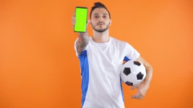 Futbol fanatiği beyaz mavi gömlekli, yeşil ekran reklam konsepti olan akıllı telefonu gösteriyor.