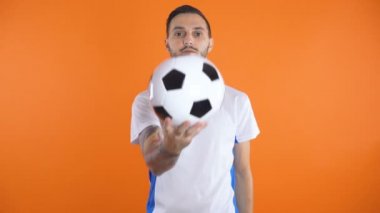Beyaz mavi tişörtlü genç futbolcu elinde futbol topuyla kameranın önünde ciddi bir yüz ifadesiyle.