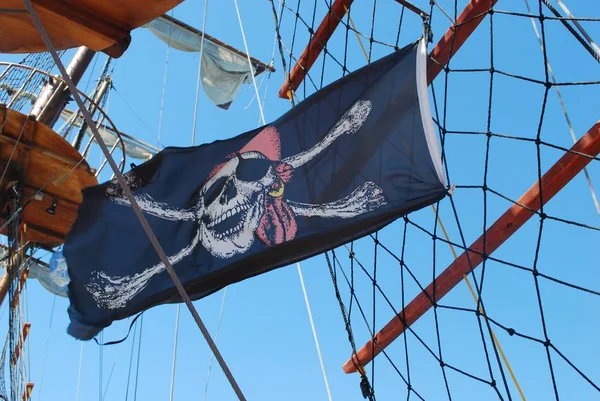 Masts and a rigging of sail vessel (yachts) against a blue sky. Joke pirate black flag with skull and crossbones fluttering in the wind