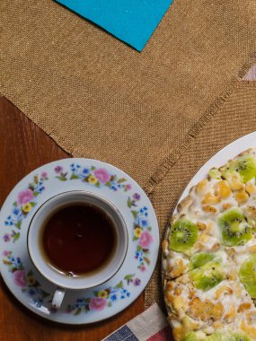 Yanında bir daire ve siyah çay damalı peçete üzerinde beyaz bir tabak bisküvi, kivi dilimleri ile dekore edilmiş yapılmış bir kek yatıyor.