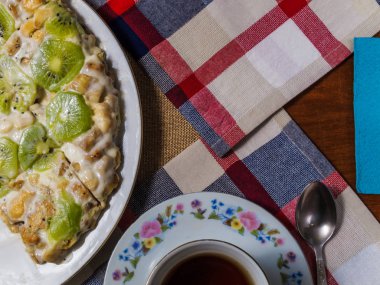 Yanında bir daire ve siyah çay damalı peçete üzerinde beyaz bir tabak bisküvi, kivi dilimleri ile dekore edilmiş yapılmış bir kek yatıyor.