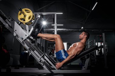 Modern spor salonunda bacak ütüleme makinesi üzerinde çalışan şortlu yakışıklı, kaslı, vahşi Avrupalı bir adamın motivasyon portresi. Gece egzersizi konsepti.