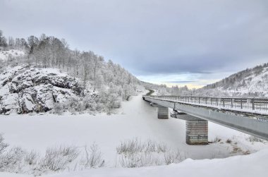 Karayolu Köprüsü aracılığıyla karlı dağlar - güzel kış alacakaranlık manzara kayalıklarla ile. Huş ağacı ve çam ormanı ile hoarfrost tepelerin eteklerinde, uzak yol arabalar kış yolcuları ile kaplı