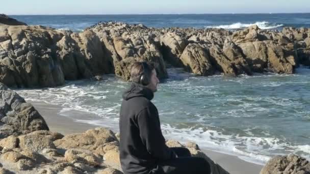 Hombres en auriculares practicando yoga en el océano Pacífico costa rocosa con magníficas olas — Vídeos de Stock