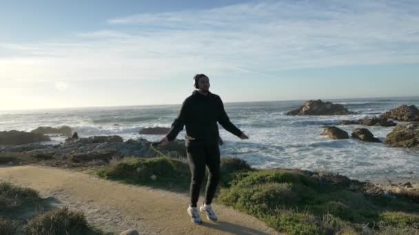 Muži se sluchátky Přeskakování školení na pobřeží Pacifiku s nádhernými vlnami za slunečného dne cvičení je zdravé fitness tělo — Stock video