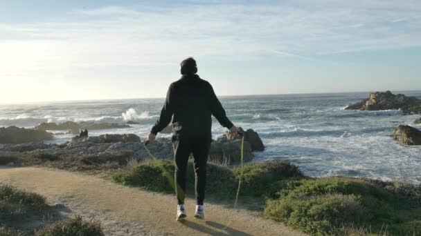 Homens com fones de ouvido Saltar o treinamento na costa pacífica com ondas magníficas no dia ensolarado exercitar-se é corpo de fitness saudável — Vídeo de Stock