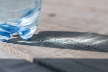 Tahta bir yüzey üzerinde maden suyu bulunan plastik bir şişe duruyor. Güneş ışınları suda parlıyor ve tahtaların üzerinde soyut bir desen oluşturuyor..