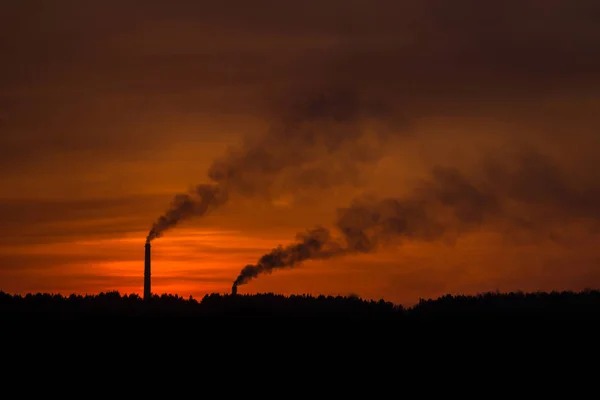 Silhouette of smoking factory pipes against the sunset landscape. Two pipes emit thick smoke.