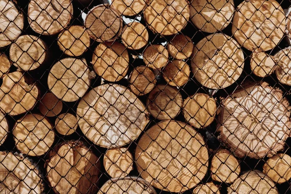 The sawn logs prepared for the production of firewood lie in rows on top of each other behind a metal grid. The wood lies behind the chain-link mesh.