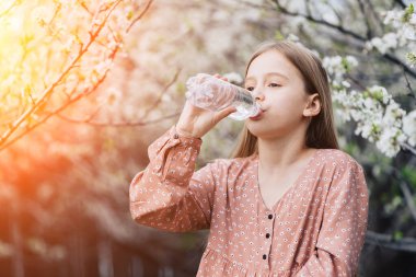 Küçük bir kız bahçedeki çiçekli bir ağacın yanındaki plastik şişeden temiz su içiyor..