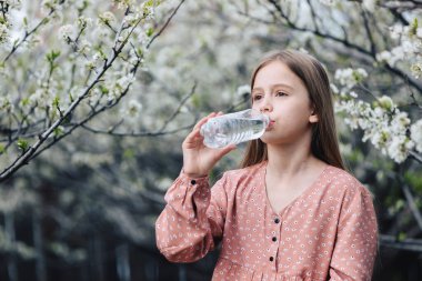 Küçük bir kız bahçedeki çiçekli bir ağacın yanındaki plastik şişeden temiz su içiyor..