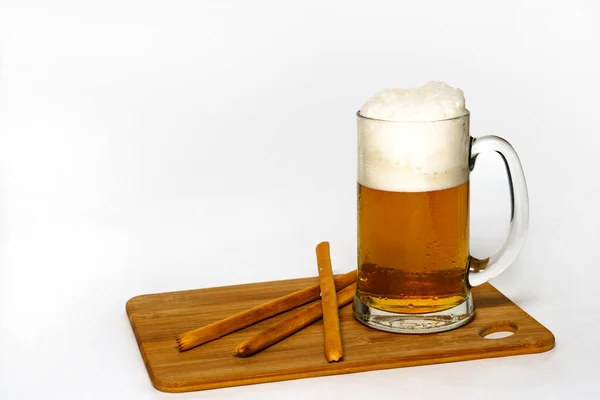 Bread sticks and misted mug of beer on a wooden cutting board. Everything is on a white background. Foam and drops.