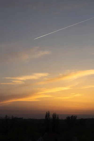 Üzerinde uçan bir uçağın izi olan karanlık bir şehrin arka planında sarı-mavi gün batımı.