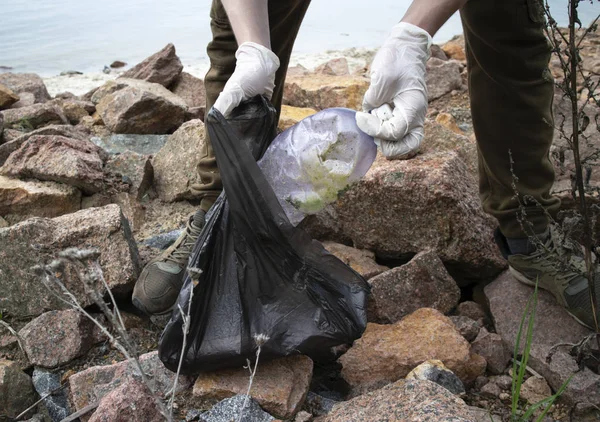 Young man collects plastic garbage in a garbage bag on the rocky beach full of rigid plastic bottles and other garbage washed out on coast of the Kiev Sea. (Volunteering concept)